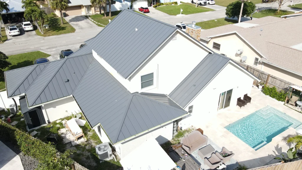 Aerial drone view of a modern two-story white house featuring a newly installed matte gray standing seam metal roof, complete with a backyard swimming pool and patio lounge area.