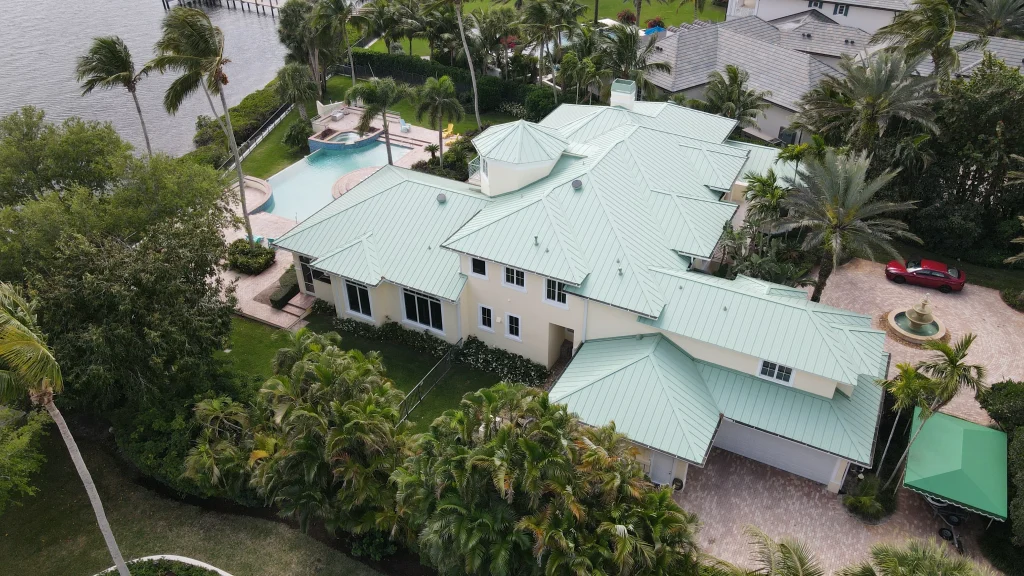 Aerial view of a massive Mediterranean-style waterfront estate featuring a striking mint green standing seam metal roof installed by Ace Pro Roofing.