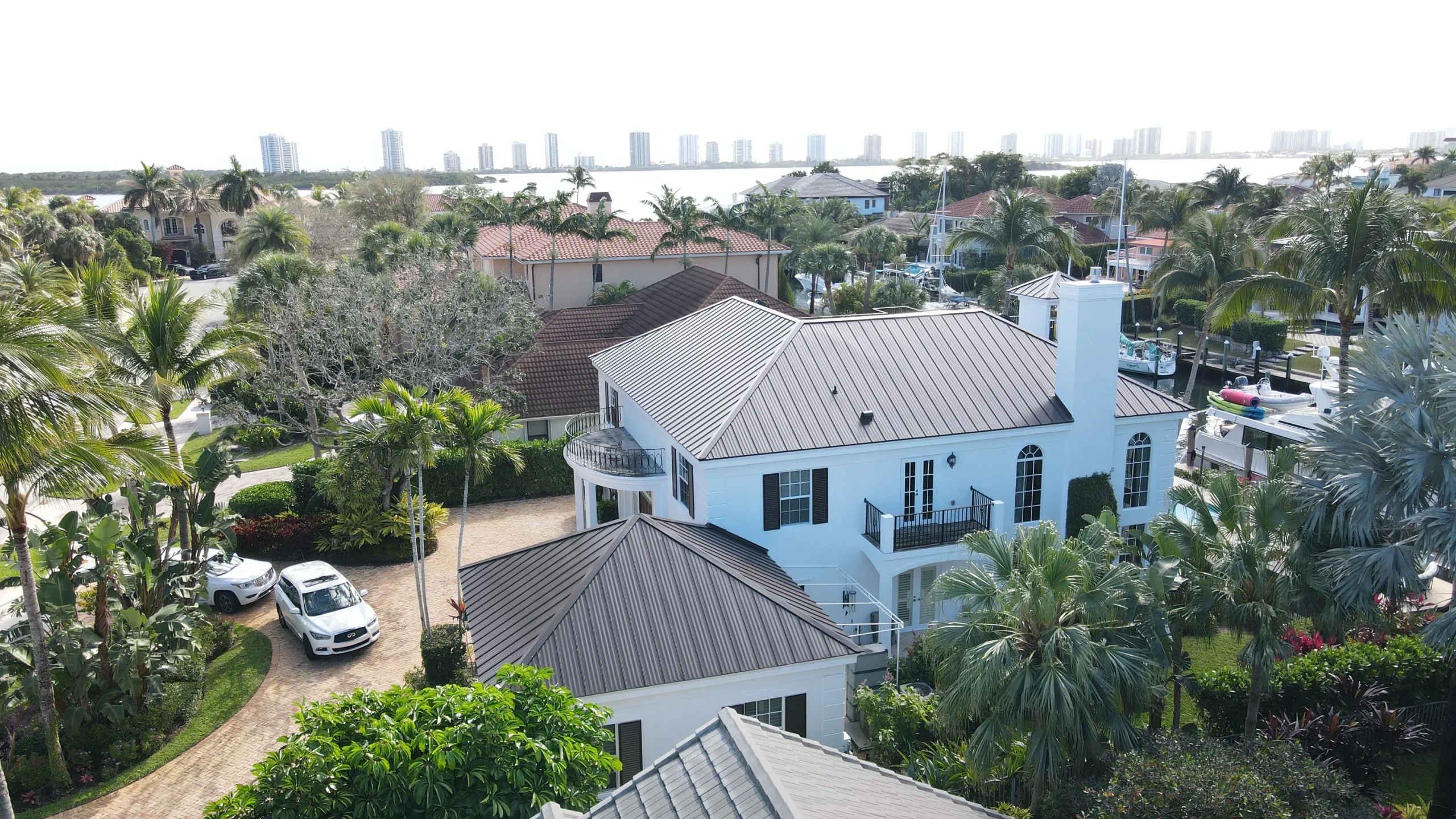Scenic drone view of a white multi-story coastal mansion with a newly installed dark metal roof by Ace Pro Roofing, with the Palm Beach area Intracoastal Waterway in the background.