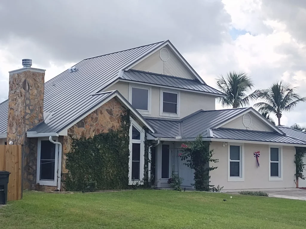 Front exterior view of a two-story residential home with a newly installed charcoal gray standing seam metal roof, stone chimney accent, and lush green lawn by Ace Pro Roofing.
